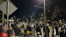 Скандал з ТЦК у Львові. Нічний горизонтальний знімок великого натовпу переважно молодих людей на міській вулиці. Вони стоять і пересуваються під тьмяним світлом ліхтарів і вікон багатоповерхівок на фоні. На передньому плані виділяються: чоловік у синій шапці, чоловік у камуфляжі, хлопець із жовтим рюкзаком Glovo, дівчина в білій кофті та хлопець у темній футболці. Праворуч натовп змішується. На задньому плані зліва припарковані фургон і вантажівка, частково закриті людьми. Високі ліхтарні стовпи з проводами перетинають небо. Справа видно червоне світло. Типовий житловий район із деревами. Атмосфера жвава й хаотична. Низький ракурс підкреслює масштаб зібрання.