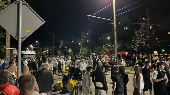 Скандал з ТЦК у Львові. Нічний горизонтальний знімок великого натовпу переважно молодих людей на міській вулиці. Вони стоять і пересуваються під тьмяним світлом ліхтарів і вікон багатоповерхівок на фоні. На передньому плані виділяються: чоловік у синій шапці, чоловік у камуфляжі, хлопець із жовтим рюкзаком Glovo, дівчина в білій кофті та хлопець у темній футболці. Праворуч натовп змішується. На задньому плані зліва припарковані фургон і вантажівка, частково закриті людьми. Високі ліхтарні стовпи з проводами перетинають небо. Справа видно червоне світло. Типовий житловий район із деревами. Атмосфера жвава й хаотична. Низький ракурс підкреслює масштаб зібрання.