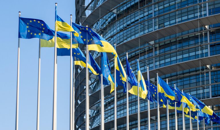 Ряд прапорів Європейського Союзу та України майорять на флагштоках на тлі сучасної скляної будівлі Європейського парламенту у Страсбурзі під ясним синім небом. Тимчасовий захист для українців у ЄС.