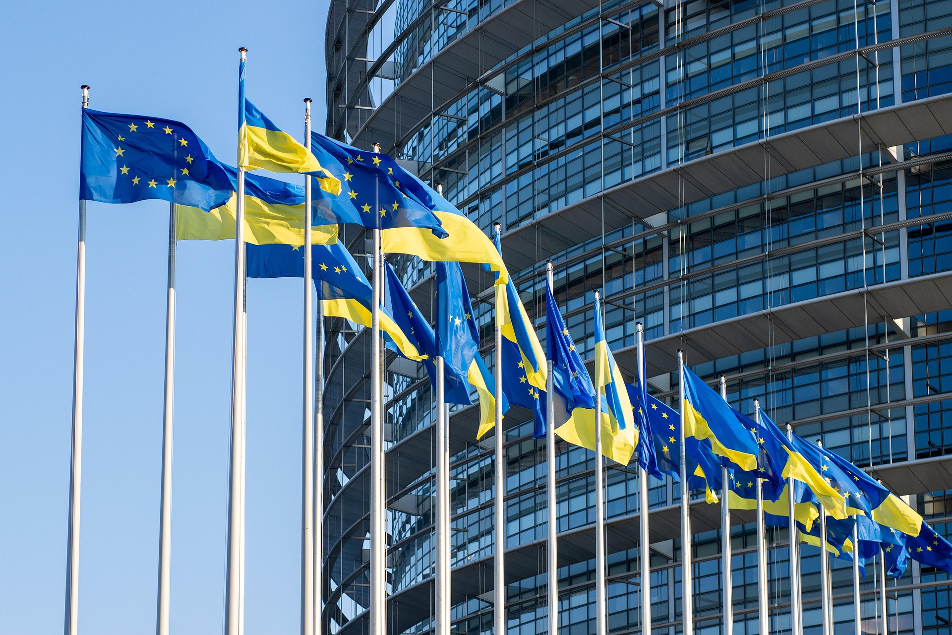 Ряд прапорів Європейського Союзу та України майорять на флагштоках на тлі сучасної скляної будівлі Європейського парламенту у Страсбурзі під ясним синім небом. Тимчасовий захист для українців у ЄС.