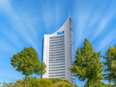 Снимок, запечатлевший небоскреб MDR (Mitteldeutscher Rundfunk) в Лейпциге, Германия, снятый снизу вверх. Справа и слева на переднем плане видны две пышные зеленые кроны деревьев, обрамляющие здание. Монументальный фасад высотного здания, выполненный из серого гранита и стекла, устремляется в яркое голубое небо. На изогнутой части фасада виден синий логотип MDR. Небоскреб имеет характерную заостренную форму и панорамное остекление на верхних этажах, а также антенну на самой вершине. Небо пронизано белыми лучами, расходящимися от центра. Снимок сделан в солнечный день с естественным освещением. Лейпциг обгоняет Штутгарт