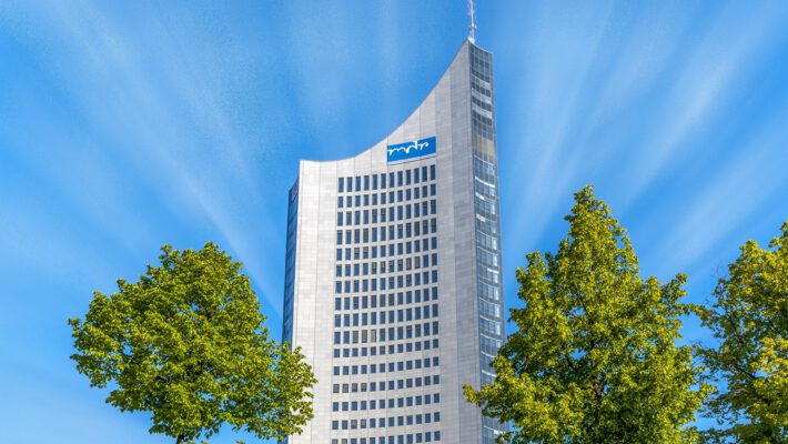 Снимок, запечатлевший небоскреб MDR (Mitteldeutscher Rundfunk) в Лейпциге, Германия, снятый снизу вверх. Справа и слева на переднем плане видны две пышные зеленые кроны деревьев, обрамляющие здание. Монументальный фасад высотного здания, выполненный из серого гранита и стекла, устремляется в яркое голубое небо. На изогнутой части фасада виден синий логотип MDR. Небоскреб имеет характерную заостренную форму и панорамное остекление на верхних этажах, а также антенну на самой вершине. Небо пронизано белыми лучами, расходящимися от центра. Снимок сделан в солнечный день с естественным освещением. Лейпциг обгоняет Штутгарт