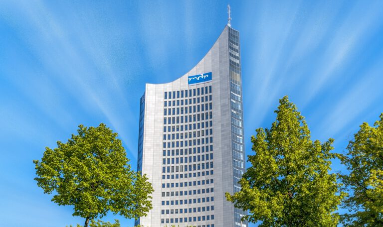 Снимок, запечатлевший небоскреб MDR (Mitteldeutscher Rundfunk) в Лейпциге, Германия, снятый снизу вверх. Справа и слева на переднем плане видны две пышные зеленые кроны деревьев, обрамляющие здание. Монументальный фасад высотного здания, выполненный из серого гранита и стекла, устремляется в яркое голубое небо. На изогнутой части фасада виден синий логотип MDR. Небоскреб имеет характерную заостренную форму и панорамное остекление на верхних этажах, а также антенну на самой вершине. Небо пронизано белыми лучами, расходящимися от центра. Снимок сделан в солнечный день с естественным освещением. Лейпциг обгоняет Штутгарт
