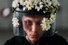 Обмеження для ухилянтів. Портрет великим планом з рівнем очей демонструє татуйованого чоловіка середнього віку, який одягнений у чорну кепку та прозору маску для обличчя з прорізами, прикрашену квітами, та дивиться на глядача. Вона дивиться на глядача прямо, трохи підвівши брови. Обличчя трохи засмагле, з довгим, гострим носом і невеликим заглибленням у щіках. Очі чоловіка сіро-блакитні. Чорний козирок закриває лоб і верхівку. По краях, попереду та по боках козирка є білі та жовтуваті квіти. Вони виглядають як ромашки. По боках обличчя чоловіка, квіти, застібка та кінчики маски. Прозора пластикова маска, прикріплена до козирка та розтягнута навколо обличчя, має білі, горизонтальні, вузькі прорізи на щоках та підборідді. Одягнута чорна футболка. Татуювання на шиї та підборідді, але вони нечіткі. Фон розмитий і сірий. Освітлення природне.