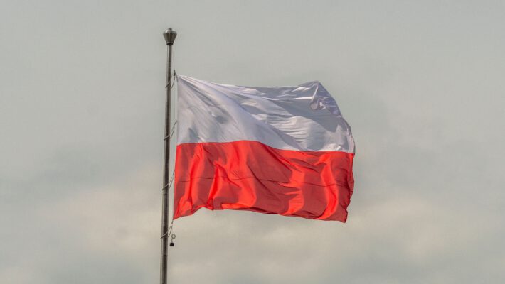 Національний прапор Республіки Польща, що складається з двох рівновеликих горизонтальних смуг — верхньої білої та нижньої червоної. Прапор зафіксований на високому металевому флагштоку і хвилюється під дією вітру. Фрагмент кадру фокусується на полотнищі, задній план — нейтральне, затягнуте світло-сірими хмарами небо.