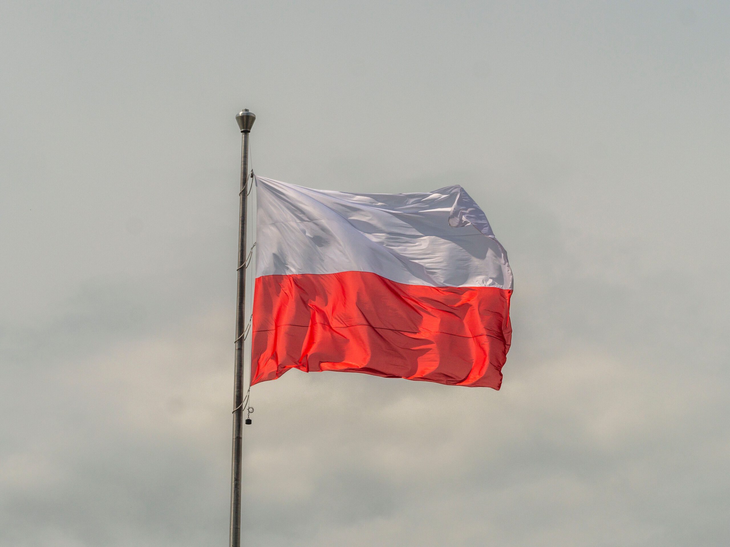 Національний прапор Республіки Польща, що складається з двох рівновеликих горизонтальних смуг — верхньої білої та нижньої червоної. Прапор зафіксований на високому металевому флагштоку і хвилюється під дією вітру. Фрагмент кадру фокусується на полотнищі, задній план — нейтральне, затягнуте світло-сірими хмарами небо.