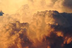 Крупный план массивных, бугристых, объемных кучево-дождевых облаков во время заката или восхода солнца. Солнечный свет окрашивает облака в насыщенный, глубокий оранжевый цвет с более светлыми золотистыми участками. Торнадо и ураганы в Германии.