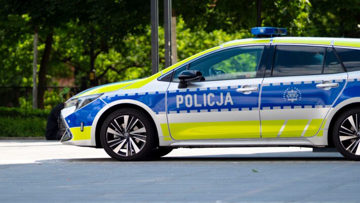Вид сбоку на современный польский патрульный полицейский автомобиль Toyota Corolla Touring Sports в новой серебристо-сине-желтой цветовой схеме. Машина с включенной синей люстрой припаркована на мощеной площади. На передней двери видна крупная надпись "POLICJA". На задней двери изображен герб полиции и надписи "POMAGAMY I CHRONIMY", "POLICJA" и номер "112". Фоном служат размытые зеленые деревья и городская инфраструктура. Снимок сделан в солнечный день. 91-летняя пенсионерка.