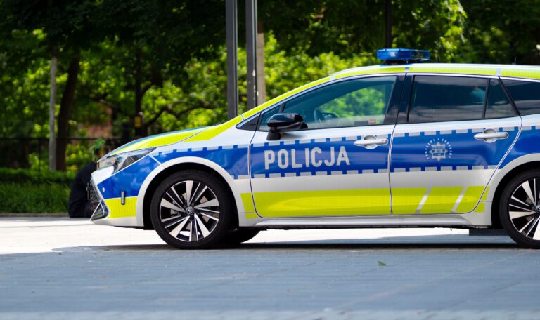 Вид сбоку на современный польский патрульный полицейский автомобиль Toyota Corolla Touring Sports в новой серебристо-сине-желтой цветовой схеме. Машина с включенной синей люстрой припаркована на мощеной площади. На передней двери видна крупная надпись "POLICJA". На задней двери изображен герб полиции и надписи "POMAGAMY I CHRONIMY", "POLICJA" и номер "112". Фоном служат размытые зеленые деревья и городская инфраструктура. Снимок сделан в солнечный день. 91-летняя пенсионерка.