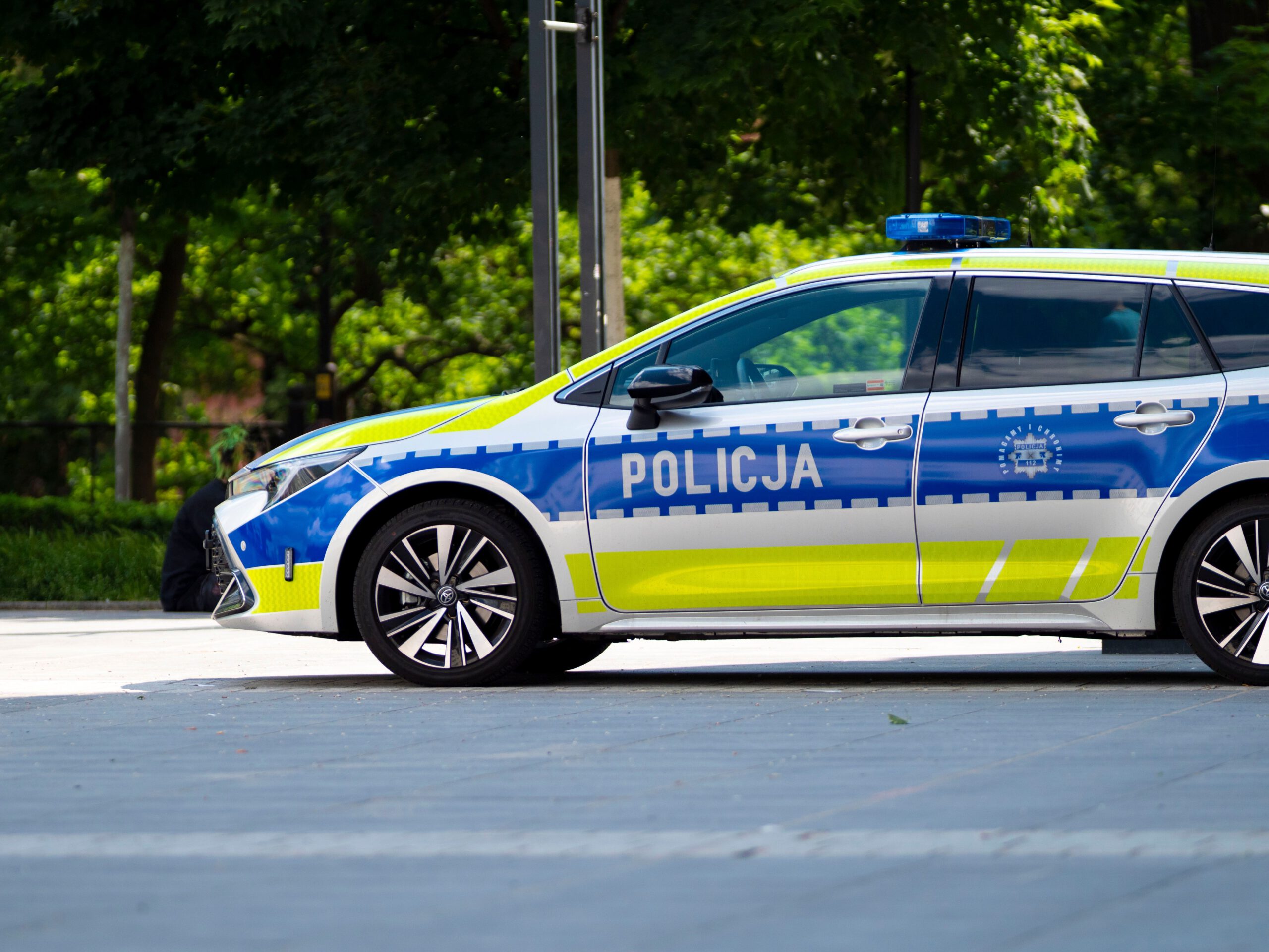 Вид сбоку на современный польский патрульный полицейский автомобиль Toyota Corolla Touring Sports в новой серебристо-сине-желтой цветовой схеме. Машина с включенной синей люстрой припаркована на мощеной площади. На передней двери видна крупная надпись "POLICJA". На задней двери изображен герб полиции и надписи "POMAGAMY I CHRONIMY", "POLICJA" и номер "112". Фоном служат размытые зеленые деревья и городская инфраструктура. Снимок сделан в солнечный день. 91-летняя пенсионерка. Вид сбоку на современный польский патрульный полицейский автомобиль Toyota Corolla Touring Sports в новой серебристо-сине-желтой цветовой схеме. Машина с включенной синей люстрой припаркована на мощеной площади. На передней двери видна крупная надпись "POLICJA". На задней двери изображен герб полиции и надписи "POMAGAMY I CHRONIMY", "POLICJA" и номер "112". Фоном служат размытые зеленые деревья и городская инфраструктура. Снимок сделан в солнечный день. 91-летняя пенсионерка.