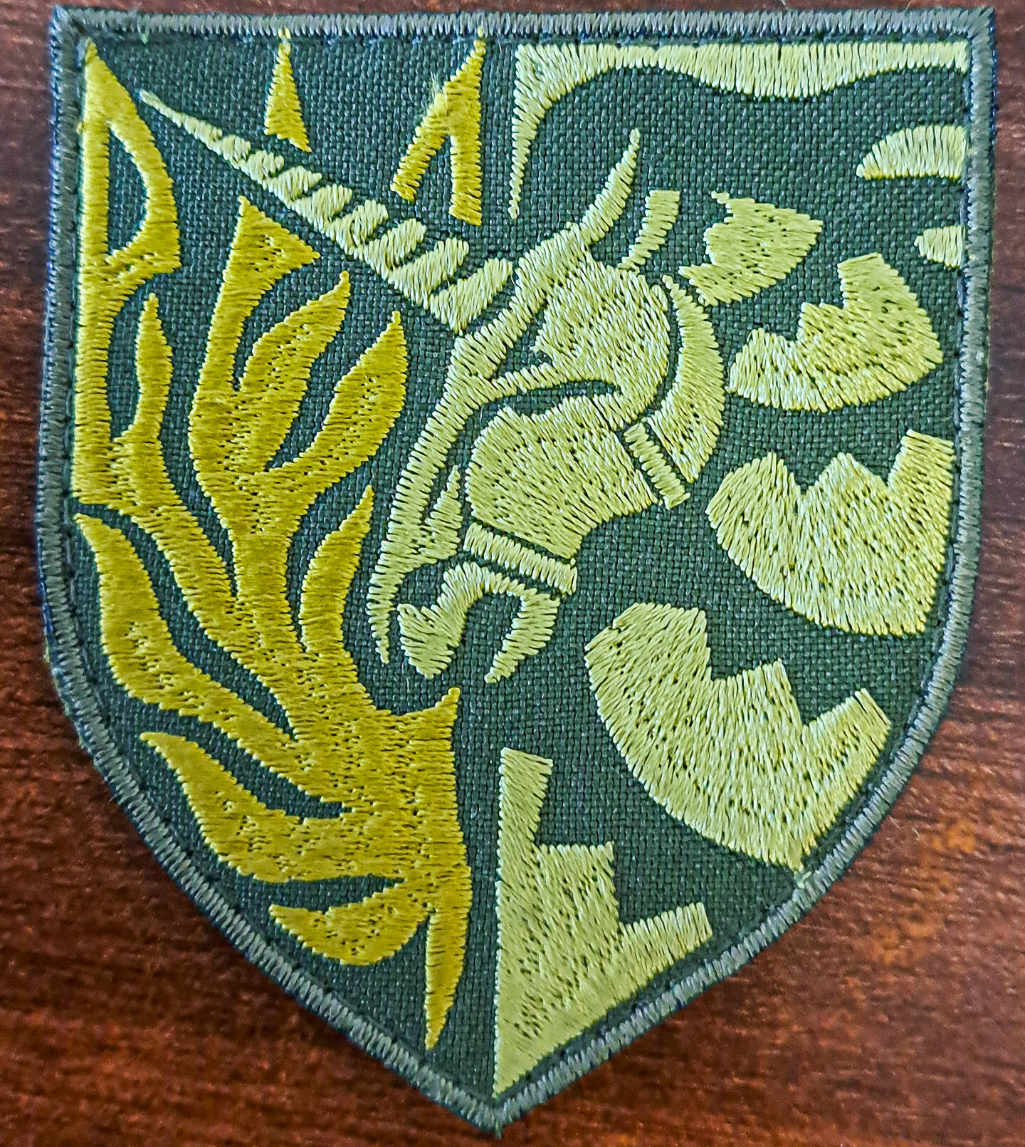 Ein gesticktes Wappen in Form eines Schildes auf dunkelgrünem Stoff. Es zeigt goldgelbe und hellgrüne Pflanzenmotive und ist mit einer dekorativen Umrandung versehen.