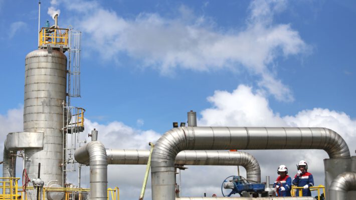 Запаси газу в сховищах ФРН. На тлі блакитного неба з білими хмарами розташований промисловий об'єкт із великою циліндричною металевою вежею та жовтими металоконструкціями. Праворуч на передньому плані видно мережу звивистих сталевих труб, а на платформі стоять двоє робітників у захисних касках та спецодязі.