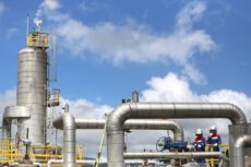 Запасы газа в хранилищах ФРГ. Промышленная башенная установка и сеть стальных труб на электростанции, рядом с которыми работают инженеры в защитных касках и сине-красной спецодежде. Запасы газа в хранилищах ФРГ. Промышленная башенная установка и сеть стальных труб на электростанции, рядом с которыми работают инженеры в защитных касках и сине-красной спецодежде.
