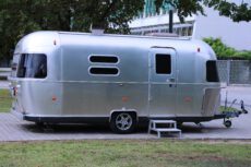 На фото изображен классический компактный автодом (кемпер) марки Airstream с узнаваемым закругленным кузовом из полированного серебристого алюминия. Трейлер припаркован на вымощенной плиточным покрытием площадке рядом с зеленой газоном и деревьями. У входа установлена небольшая серая ступенька для удобства, на окнах видны темные тонированные стекла, а спереди расположен металлический сцепной механизм для транспортировки. Илон Маск живет в автотрейлере.