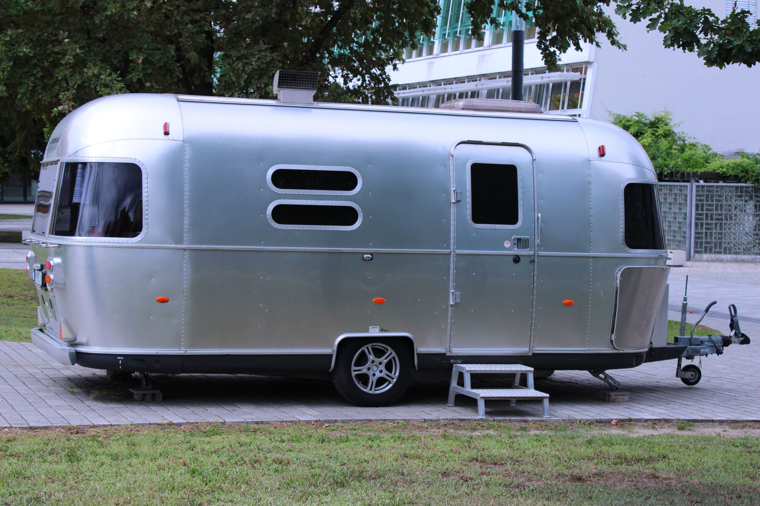На фото изображен классический компактный автодом (кемпер) марки Airstream с узнаваемым закругленным кузовом из полированного серебристого алюминия. Трейлер припаркован на вымощенной плиточным покрытием площадке рядом с зеленой газоном и деревьями. У входа установлена небольшая серая ступенька для удобства, на окнах видны темные тонированные стекла, а спереди расположен металлический сцепной механизм для транспортировки. Илон Маск живет в автотрейлере.