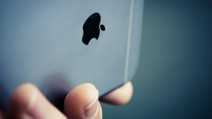 Верхня частина сірої задньої панелі телефону Apple, що тримається горизонтально, знаходиться у фокусі. Зліва внизу її закриває верхня частина безіменного та мізинця лівої руки з нігтями, які тримають пристрій. В центрі задньої панелі розташоване надкушене яблуко. Зверху над яблуком на задній панелі, і ще вище, де вона стикається з торцем, проходять дві горизонтальні смуги. На тлі нерівномірно затемнене розмите узлісся в сірих відтінках. iCloud+ безкоштовно Apple TV Arcade Україна.