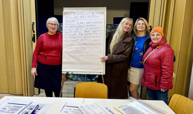 Викладач німецької мови у червоній кофті та окулярах стоїть поруч із фліпчартом, на якому вручну написано навчальний текст про погоду та цитату Тоніно Гуерри німецькою мовою. Поруч із нею посміхаються три учасниці заняття, а на передньому плані розкладено навчальні матеріали.