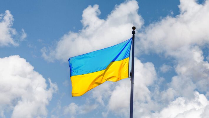 На фото зображено державний прапор України, який майорить на високому флагштоку. Полотнище прапора складається з двох рівних горизонтальних смуг: синьої зверху та жовтої знизу. Флагтсток розташований праворуч і йде вертикально вгору. На тлі видно яскраво-блакитне небо, покрите легкими, пухнастими білими хмарами. Освітлення денне, природне, що підкреслює яскраві кольори прапора. Підтримка вступу України в ЄС у Німеччині.