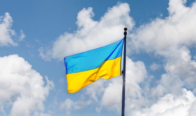 На фото зображено державний прапор України, який майорить на високому флагштоку. Полотнище прапора складається з двох рівних горизонтальних смуг: синьої зверху та жовтої знизу. Флагтсток розташований праворуч і йде вертикально вгору. На тлі видно яскраво-блакитне небо, покрите легкими, пухнастими білими хмарами. Освітлення денне, природне, що підкреслює яскраві кольори прапора. Підтримка вступу України в ЄС у Німеччині.