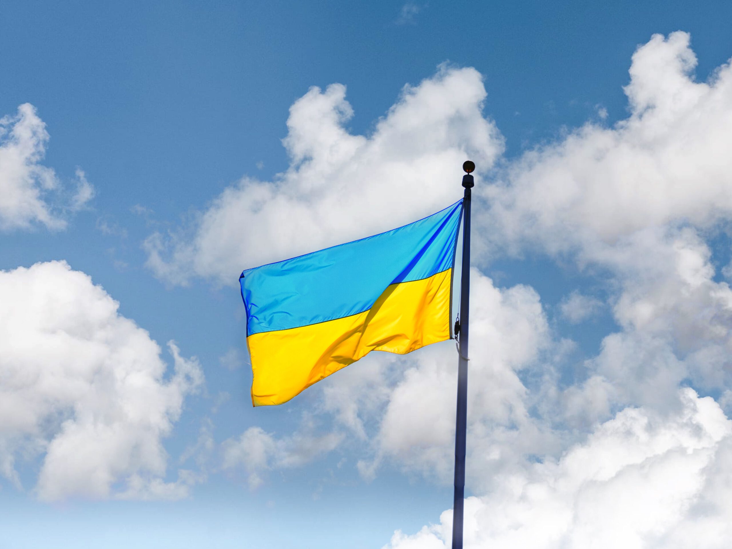 На фото зображено державний прапор України, який майорить на високому флагштоку. Полотнище прапора складається з двох рівних горизонтальних смуг: синьої зверху та жовтої знизу. Флагтсток розташований праворуч і йде вертикально вгору. На тлі видно яскраво-блакитне небо, покрите легкими, пухнастими білими хмарами. Освітлення денне, природне, що підкреслює яскраві кольори прапора. Підтримка вступу України в ЄС у Німеччині.